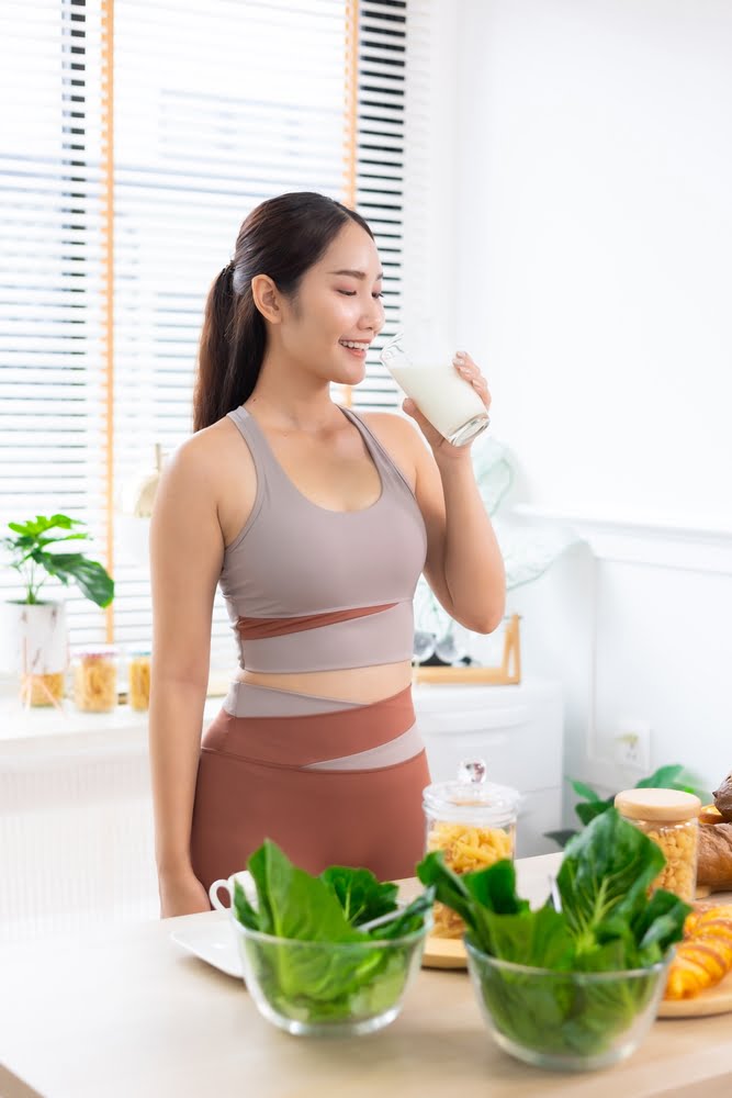 A woman in athletic wear enjoying a glass of milk in a bright kitchen, with fresh vegetables and healthy food on the counter in front of her - Chiropractor Brunswick GA A woman in athletic wear enjoying a glass of milk in a bright kitchen, with fresh vegetables and healthy food on the counter in front of her - Chiropractor Brunswick GA